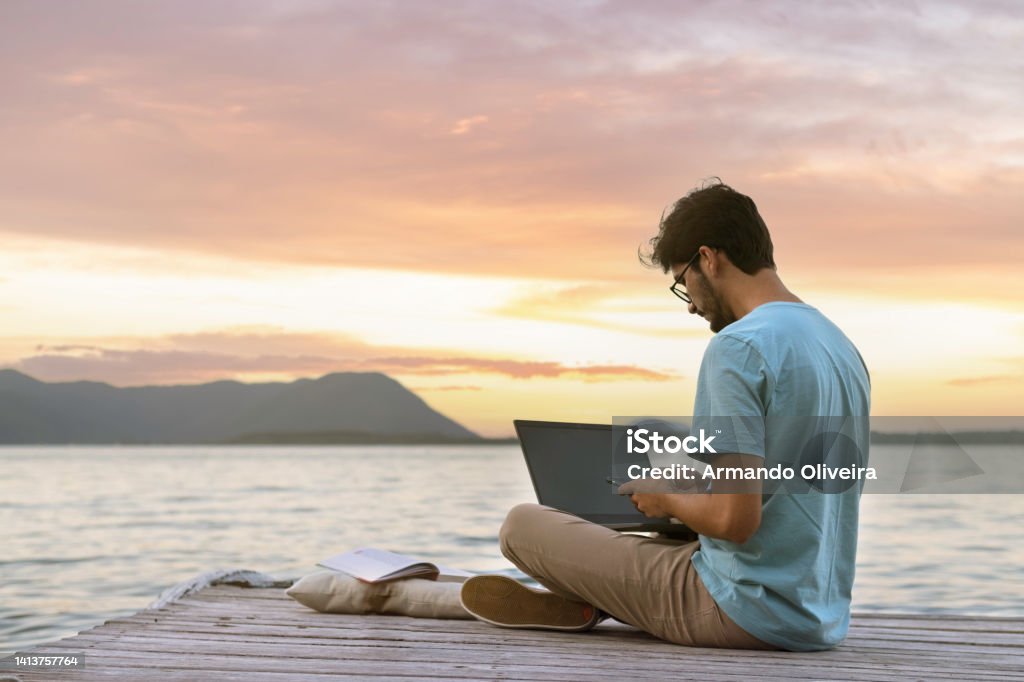 young digital nomad man sitting on wooden pier at sea working on internet remotely at sunset - Traveling with a computer - Online dream job concept